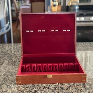 May include: A wooden silverware storage box with a red velvet interior. The box is open, revealing compartments for utensils. The exterior is a warm brown with brass accents on the corners and a gold clasp.