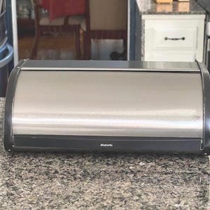 May include: A rectangular bread box with a brushed stainless steel finish and black side panels. The front features a roll-top door with a black handle strip that displays the white Brabantia brand logo centered on the lower edge of the unit.