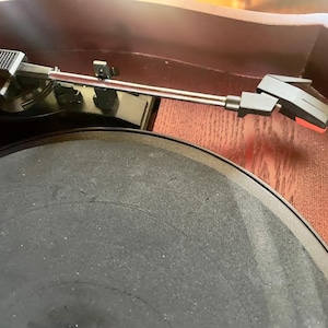 May include: Close-up of a vintage record player. The turntable features a black platter and a silver tone arm with a black headshell. The wooden cabinet has a dark brown finish.