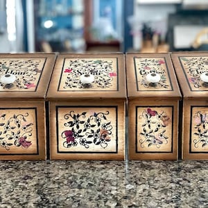 Könnte beinhalten: Vier Küchenbehälter aus Holz mit Deckeln und weißen Knöpfen. Die Deckel sind mit "Flour", "Sugar", "Coffee" und "Tea" beschriftet. Jeder Behälter hat ein dekoratives Blumen- und Schmetterlingsdesign.