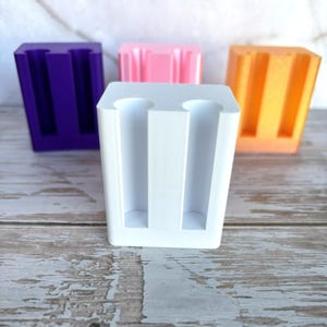 May include: A collection of four rectangular plastic holders, each featuring two vertical cylindrical cutouts. The holders are arranged in a group and come in solid white, purple, light pink, and orange colors, with a smooth, matte finish.