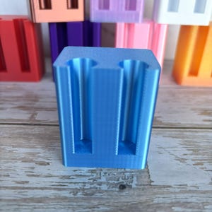 May include: A bright blue 3D printed rectangular holder featuring two vertical cylindrical cutouts on the front face. The object has a smooth, layered texture and is positioned in front of several other similar holders in various colors.