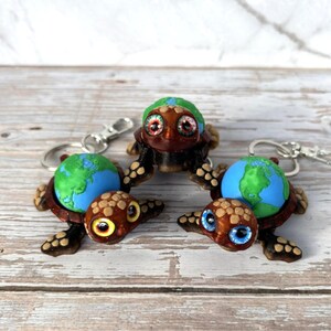 May include: Keychains featuring turtle figurines with globe shells. The turtles have brown bodies, patterned shells, and expressive eyes. The globes are blue and green, depicting the Earth. Each keychain has a silver clasp.