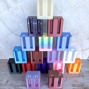 May include: A collection of small, rectangular, 3D-printed holders arranged in a pyramid shape. Each holder features two vertical cylindrical slots. The items come in a variety of solid, metallic, and gradient colors, including black, red, blue, white, orange, and purple. One holder contains a small glass vial.