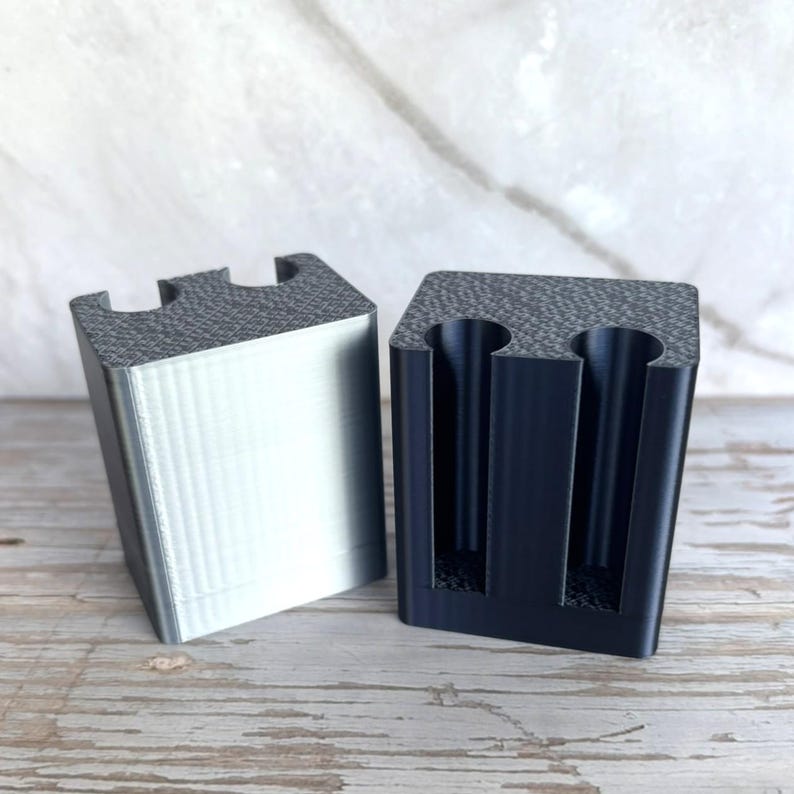 May include: Two rectangular blocks with a textured top surface and two vertical cylindrical cutouts. One block is metallic silver with a smooth finish, while the other is matte black. Both items feature a flat base and a structured, geometric design.