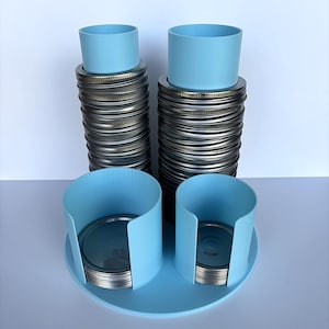 May include: A collection of canning jar lids and blue accessories. Two stacks of silver lids are topped with light blue cylindrical holders. Two additional blue holders sit on a matching blue tray, each holding a stack of lids.