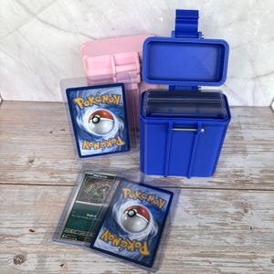 May include: A bright blue plastic storage case with an open lid, containing several clear protective card sleeves. Two additional Pokémon trading cards in clear sleeves are positioned in front of the case, alongside a light pink storage case.