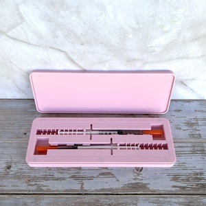 May include: A light pink case, open to reveal two syringes with orange caps. The case is rectangular with a matching lid. The syringes are black and clear, with measurements visible. The background is a light wood surface.