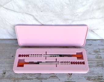 May include: A light pink case, open to reveal two syringes with orange caps. The case is rectangular with a matching lid. The syringes are black and clear, with measurements visible. The background is a light wood surface.