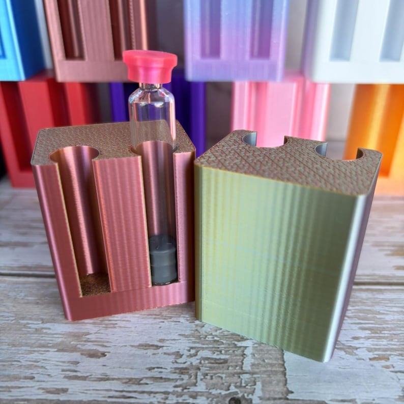 May include: Two rectangular 3D-printed holders with vertical cylindrical cutouts. The left holder is metallic rose gold and contains a clear glass vial with a pink cap and a grey stopper. The right holder features a metallic green-to-gold gradient.