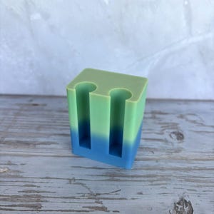 May include: A rectangular block-shaped holder featuring a smooth gradient transition from light green at the top to a vibrant blue at the base. The top surface contains two vertical cylindrical cutouts designed for holding small items upright.