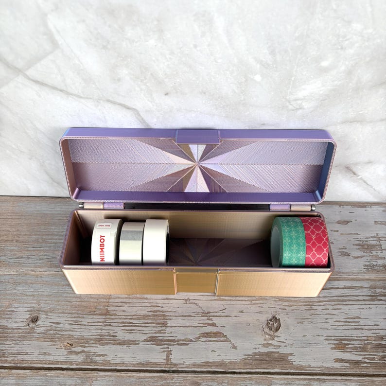 May include: An open, rectangular storage box with a hinged lid. The box is a light purple color with a geometric design on the inside of the lid. Inside are rolls of decorative tape in various colors and patterns. The box sits on a weathered wooden surface.