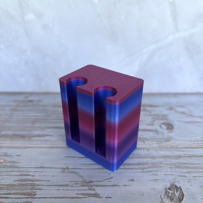 May include: A rectangular holder with two vertical cylindrical cutouts, featuring a gradient color pattern that transitions through horizontal bands of blue, purple, and magenta. The top surface has a textured finish, and the sides are smooth.