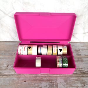 May include: A bright pink, open, plastic storage box with a hinged lid. Inside are several rolls of decorative tape in various colors and patterns. Some tape rolls have the word "NIIMBOT" printed on them.