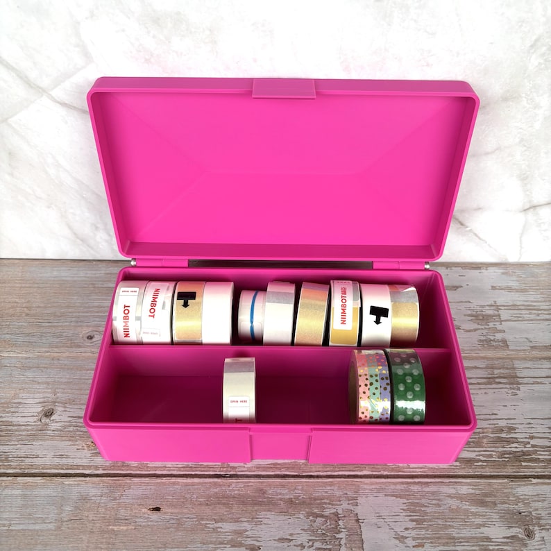 May include: A bright pink, open, plastic storage box with a hinged lid. Inside are several rolls of decorative tape in various colors and patterns. Some tape rolls have the word "NIIMBOT" printed on them.
