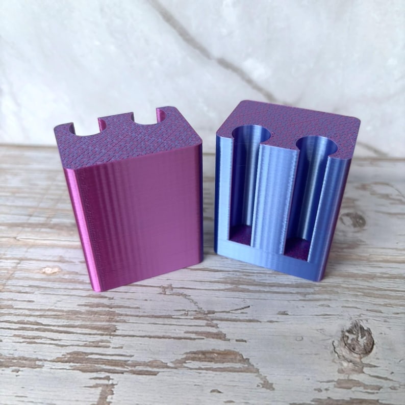 May include: Two 3D printed rectangular blocks with a color-shifting finish that transitions between purple and blue. Each block features a flat base and a top surface with two vertical cylindrical cutouts, showing a smooth, layered texture.
