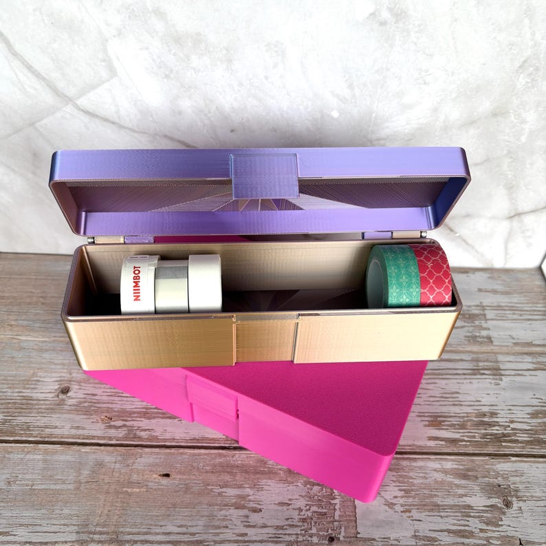 May include: Open purple and gold box with washi tape rolls inside. The box is sitting on top of a pink triangular object. The washi tape rolls include white, silver, green, and red patterned tape. The text "NIIMBOT" is visible on one of the tape rolls.