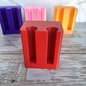 May include: A set of four rectangular holders, each featuring two vertical cylindrical cutouts. The holders are solid colors: one red in the foreground, with purple, pink, and orange holders positioned behind it in a staggered arrangement.