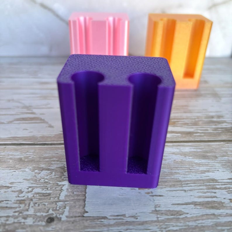 May include: A purple rectangular holder featuring two vertical cylindrical cutouts on the front face. The object has a textured surface finish and is positioned in the foreground, with a pink holder and an orange holder visible in the background.