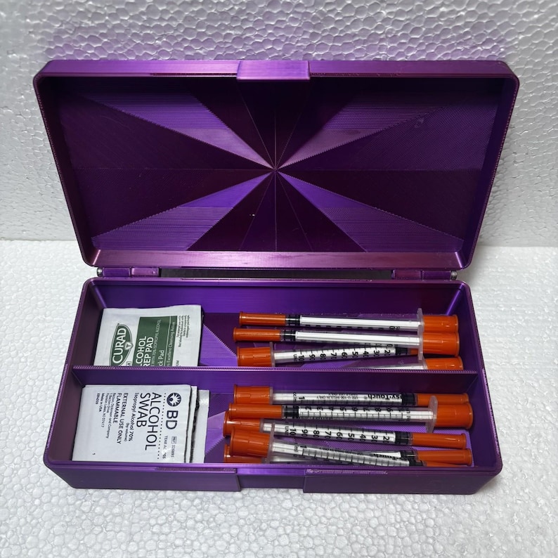 May include: A purple, open, plastic case with a starburst design on the lid. Inside are several insulin syringes with orange caps and alcohol prep pads. The case is divided into compartments for organization.
