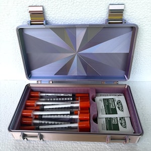 May include: Open, lavender-colored medical case with a geometric design on the inside lid. The case contains several syringes with orange caps and alcohol prep pads. The case is open, revealing its contents.