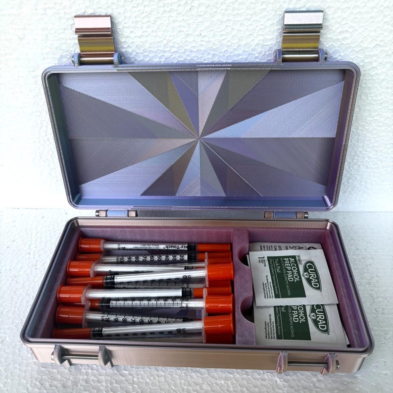 May include: Open, lavender-colored medical case with a geometric design on the inside lid. The case contains several syringes with orange caps and alcohol prep pads. The case is open, revealing its contents.