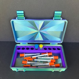 May include: An open, iridescent blue and green case with a geometric design on the lid. Inside, a purple tray holds syringes with orange caps and two small vials. The case is designed to hold medical supplies.