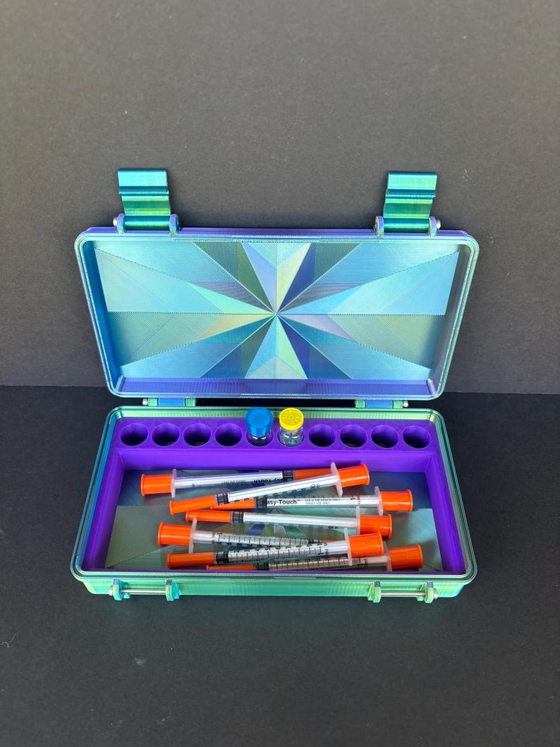 May include: An open, iridescent blue and green case with a geometric design on the lid. Inside, a purple tray holds syringes with orange caps and two small vials. The case is designed to hold medical supplies.