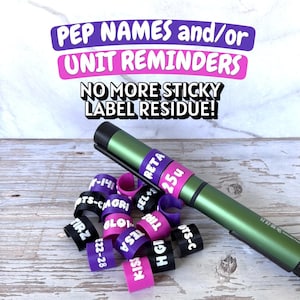 May include: A collection of small, C-shaped plastic identification rings in purple, pink, and black with white text labels like RETA, 25u, and TIRZ. A metallic green pen rests across the rings. Text above reads PEP NAMES and/or UNIT REMINDERS.