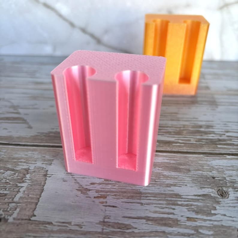 May include: A light pink, rectangular, 3D-printed plastic holder featuring two vertical cylindrical cutouts on the front face. A second, identical holder in a bright orange color is positioned slightly behind and to the right of the pink one.