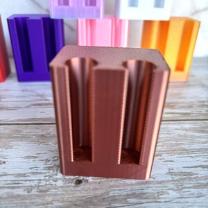 May include: A copper-colored, rectangular 3D-printed holder featuring two vertical cylindrical cutouts on the front face. The object has a smooth, matte finish and is positioned in the foreground, with several other similar holders in various colors visible behind it.