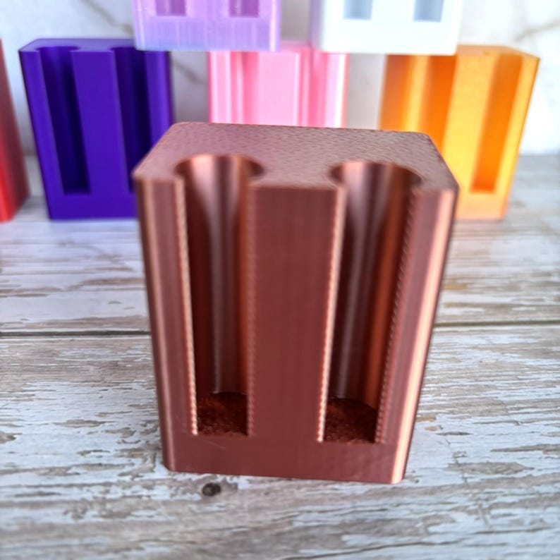 May include: A copper-colored, rectangular 3D-printed holder featuring two vertical cylindrical cutouts on the front face. The object has a smooth, matte finish and is positioned in the foreground, with several other similar holders in various colors visible behind it.