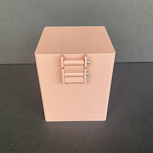 May include: A small, rectangular, blush-colored box with a hinged closure. The box has a matte finish and a simple, modern design. The hinge is made of the same material as the box and is secured with small silver screws. The box is sitting on a dark surface.