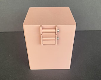 May include: A small, rectangular, blush-colored box with a hinged closure. The box has a matte finish and a simple, modern design. The hinge is made of the same material as the box and is secured with small silver screws. The box is sitting on a dark surface.