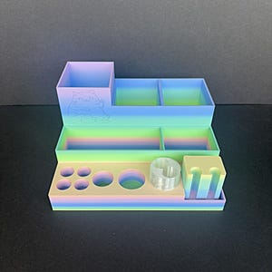 May include: A multi-tiered desk organizer in pastel rainbow colors. The organizer has various compartments, including a large square section with a cat design, several small rectangular sections, and circular and rectangular holes. A clear cylindrical object and a small square holder are also included.