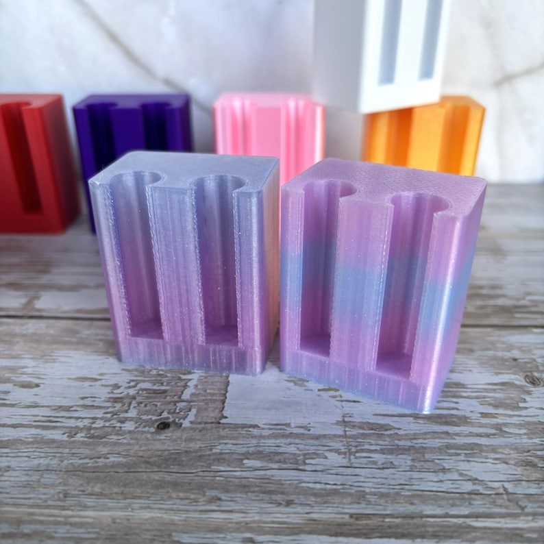 May include: Two rectangular 3D printed holders with a smooth, iridescent finish in shades of purple, pink, and blue. Each holder features two vertical cylindrical cutouts on the front face. Additional colorful holders are visible in the background.