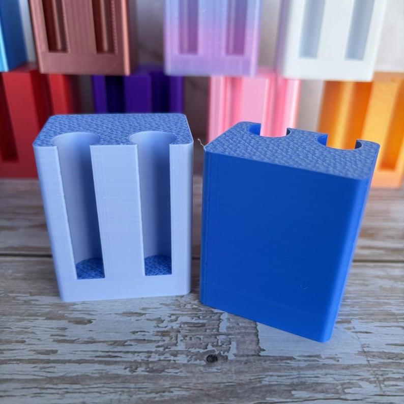 May include: Two rectangular 3D printed stands with two vertical cylindrical cutouts on the top surface. One stand is light blue and the other is a darker blue. Both feature a textured top surface and smooth sides, with additional colorful stands visible behind.