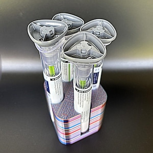 May include: A set of five medical devices with gray tops and clear tubes, each containing a green substance. They are arranged in a multi-colored holder. The devices have white labels with black text. The holder has a striped pattern.