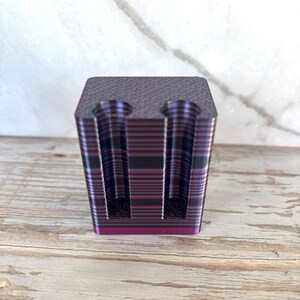 May include: A rectangular block with a textured top surface and two vertical cylindrical cutouts. The object features a gradient finish with horizontal bands of metallic purple, magenta, and dark charcoal gray colors throughout the sides.