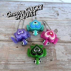 May include: Five colorful turtle-shaped keychains with heart-shaped shells. The keychains are in shades of purple, teal, pink, green, and blue. Each keychain has a small turtle head and feet. The text "Choose CLICKY or QUIET" is above the keychains.