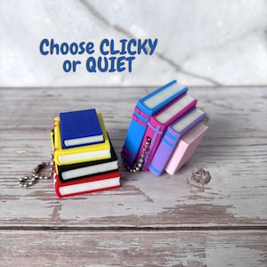 May include: Two miniature book-shaped keychains with metal ball chains. One stack features blue, yellow, black, and red covers. The other stack includes blue, pink, purple, and light pink covers. Text above reads: Choose CLICKY or QUIET.