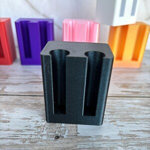 May include: A black rectangular stand featuring two vertical cylindrical cutouts on the front face. This item is part of a collection of similar stands in red, purple, pink, white, and orange, all arranged in a row behind the primary black unit.
