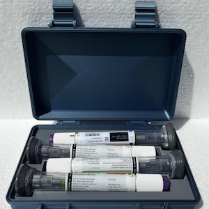 May include: A blue plastic case containing three pre-filled injection pens. Each pen is white with black and colored accents, and includes printed text. The case is open, revealing the pens neatly arranged inside.