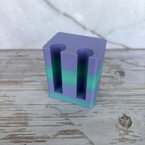May include: A rectangular block-shaped holder featuring a gradient color pattern that transitions from purple at the top to green in the middle and back to purple at the base. The object has two vertical cylindrical cutouts on the front face.