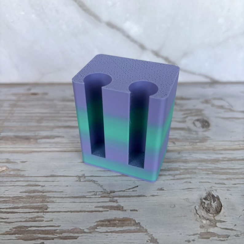 May include: A rectangular block-shaped holder featuring a gradient color pattern that transitions from purple at the top to green in the middle and back to purple at the base. The object has two vertical cylindrical cutouts on the front face.