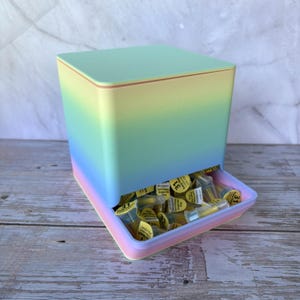 May include: A square, gradient-colored storage container transitioning from light green at the top to blue and pink at the base. The open front tray is filled with numerous small, clear plastic medical needle caps with yellow labels marked 31.