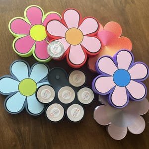 May include: A collection of colorful flower-shaped coasters in various shades of pink, orange, blue, and purple. Some coasters have a yellow or blue center. Several rolls of tape are visible in the image.