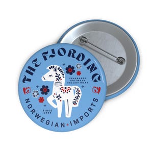 Può includere: Un bottone azzurro con la scritta "THE FJORDING NORWEGIAN + IMPORTS" che circonda un'illustrazione di cavallo bianco con accenti floreali rossi e neri. Il bottone ha anche la scritta "FRAGRANCE KNITWEAR COLLECTIBLES" e "SINCE 1988".