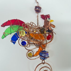 May include: A handcrafted copper rooster wind chime with colorful stained glass accents. The rooster features red, green, and blue tail feathers, orange and yellow body details, and a red comb. Decorative glass beads are incorporated throughout the design.