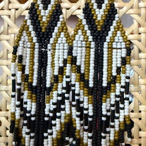 Puede incluir: Pendientes hechos a mano con cuentas y diseño geométrico. Los pendientes presentan un patrón de cuentas negras, blancas y doradas. Cuelgan de ganchos plateados. Fondo tejido.